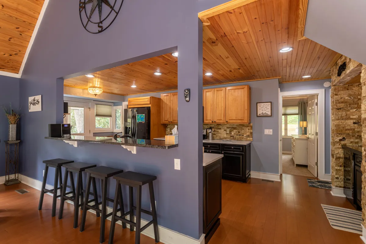 A warm and inviting interior scene showcasing a stone fireplace, bar counter with stools, and a kitchen area under a wooden ceiling. The space is set up for entertaining, relaxing by the fire, and enjoying cozy gatherings.