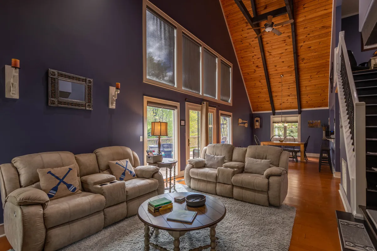 A cozy living area featuring plush sofas, a coffee table, and expansive windows looking out onto natural scenery. Exposed wooden beams add rustic charm, while the large windows allow for abundant daylight and glimpses of the outdoors.