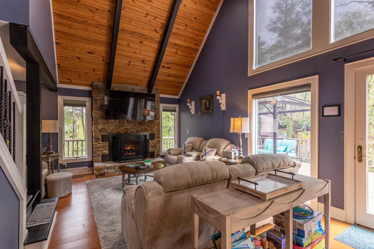 This image shows a welcoming living room centered around a striking stone fireplace, with large windows allowing sunlight to pour in and plush sofas arranged for comfort and conversation. Natural light bathes the space, creating a warm, inviting atmosphere perfect for gatherings.