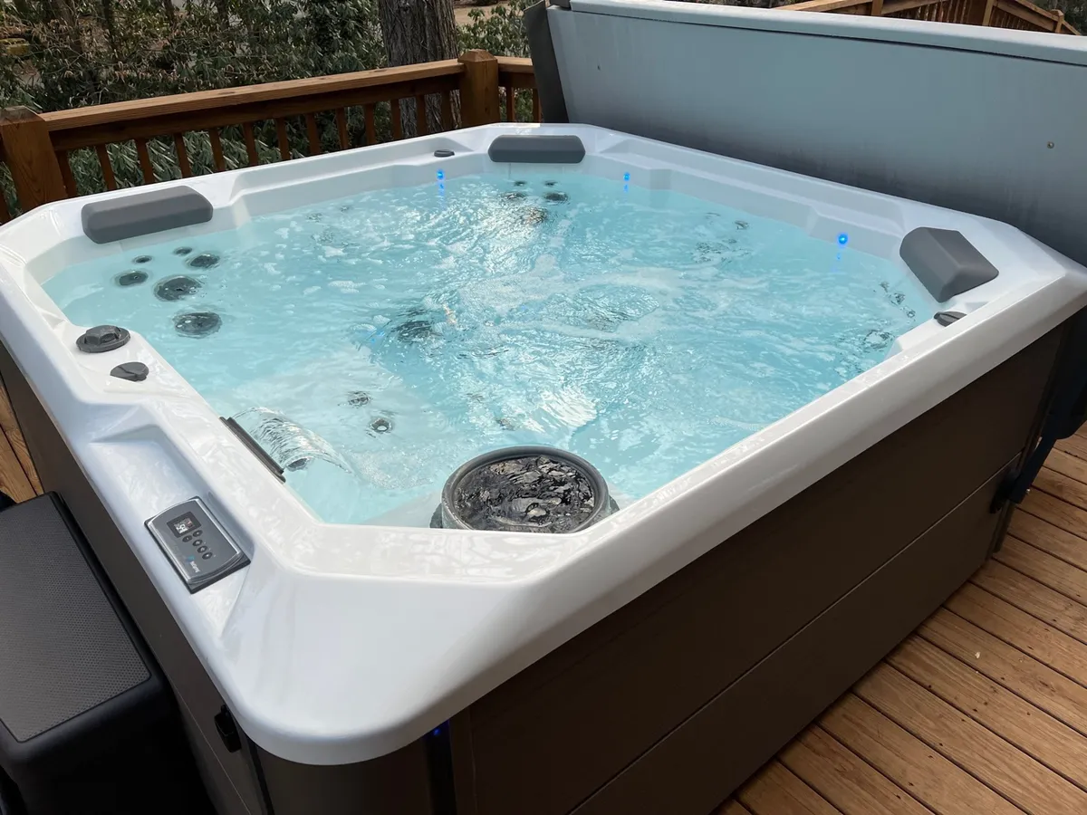 A tranquil outdoor hot tub sits on a spacious wooden deck surrounded by lush greenery, with the water's surface reflecting the morning sun. The deck provides a natural connection to the surrounding forested area, evoking a sense of relaxation and immersion in nature.