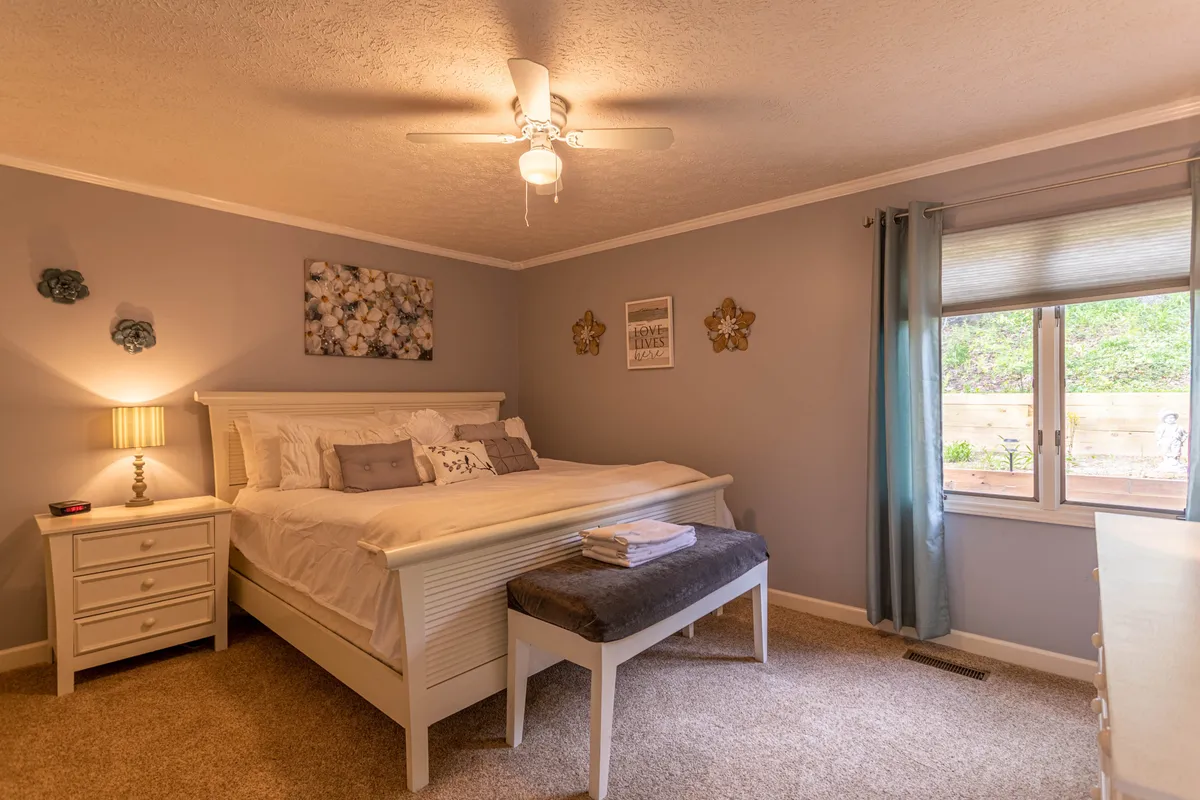 A bright bedroom with a large bed, nightstand with a lamp, decorative artwork, a window letting in natural light, and a dressing table. The scene conveys tranquility and comfort, perfect for restful sleep and quiet moments.