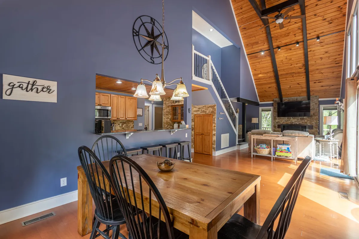 A spacious, open-concept area integrating the dining table, kitchen, and living room. The high ceiling and expansive windows amplify the natural light, while the stone fireplace and adjacent kitchen reinforce a sense of unity and warmth for shared meals and activities.