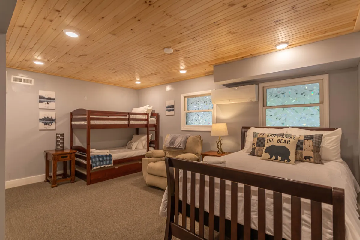 A playful bedroom designed for children, featuring a bunk bed, a single bed, an armchair with decorative pillows, and cheerful touches throughout. The space feels welcoming and fun, suitable for kids and their sleepovers.