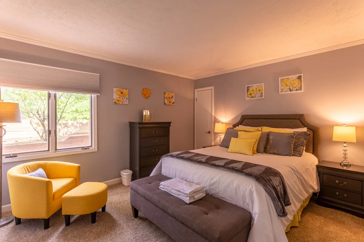 A serene bedroom with a comfortable bed, yellow armchair positioned by the window, and decorative artwork, allowing for views of nature and abundant natural light. The space feels peaceful, ideal for reading, reflecting, or enjoying a restorative break.