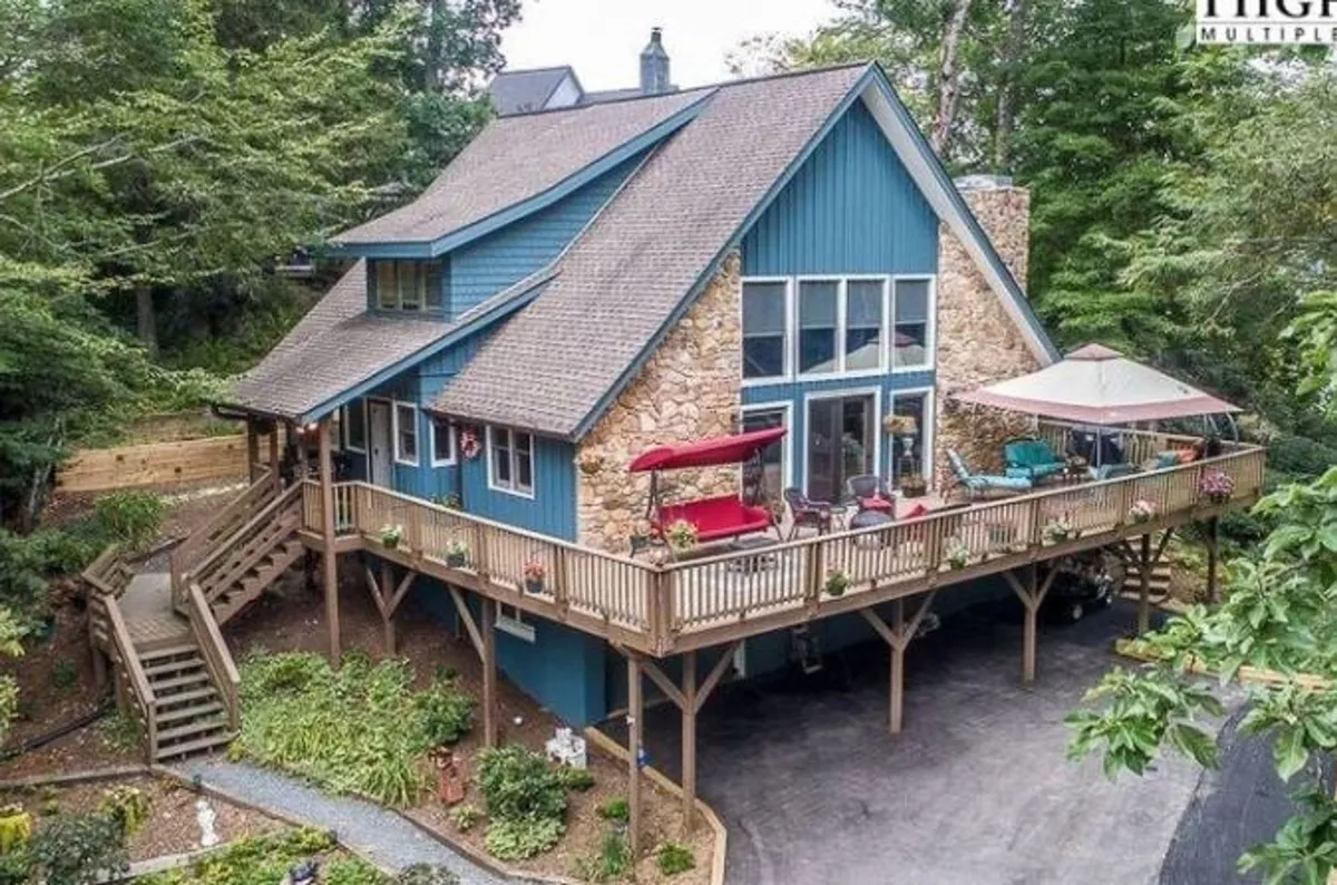 A charming blue wooden cabin stands nestled among lush greenery, its spacious stone-accented deck inviting guests to relax on comfortable seating while taking in serene mountain sunsets. The scene evokes a sense of peace and connection with nature, ideal for outdoor dining, stargazing, and unwinding in the fresh mountain air.