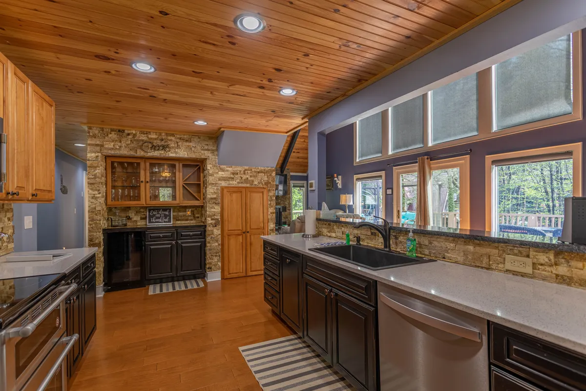 A spacious kitchen with a kitchen island, large windows letting in natural light, a wooden ceiling, and stone wall features. The space is set for group cooking, dining, and socializing, with hints of the outdoors through the windows.